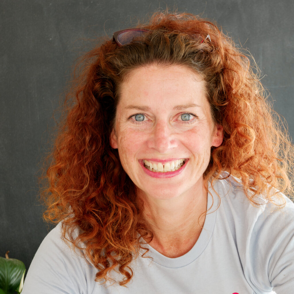 Headshot of website author. Long curly red hair, big smile, blue eyes, in a light blue tee.
